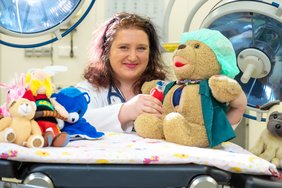 Kathrin Lindner steht im Kittel hinter einer Krankenliege auf der verschiedene Kuscheltiere sitzen. Im Hintergrund ist medizinisches Gerät zu erkennen.