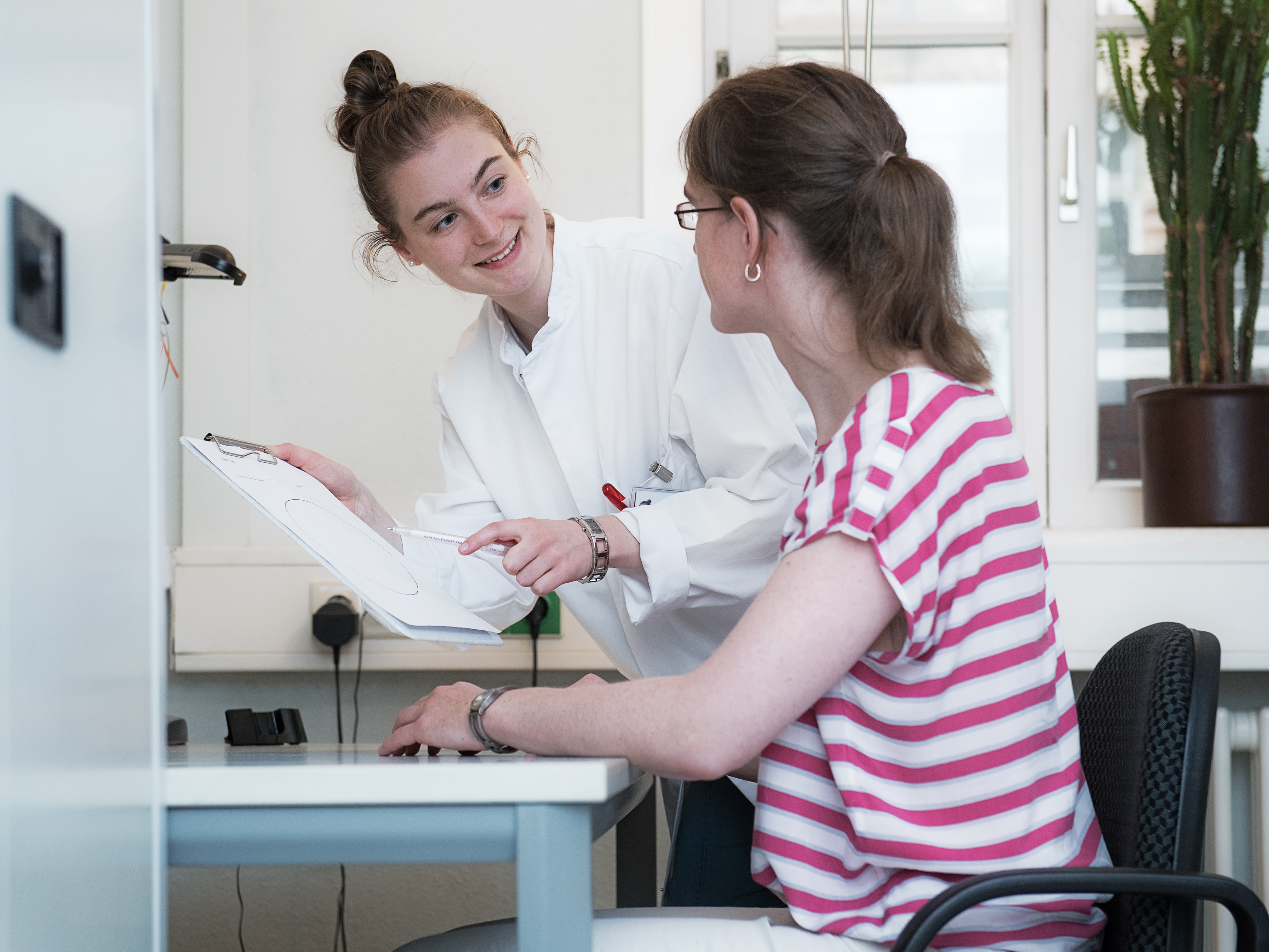 Frau im weißen Kittel mit einer Frau am Tisch, erstere hält ein Klemmbrett mit Aufgaben