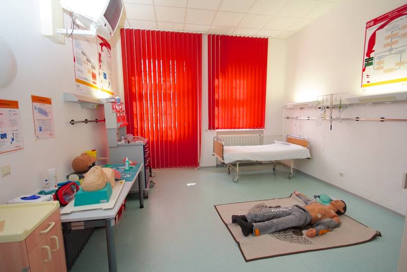 Blick in ein Übungs-Krankenhauszimmer mit Bett, Tischen, auf denen verschiedene Utensilien liegen, roten Vorhängen vor dem Fenster und einer Simulations-Puppe, die auf dem Boden liegt.