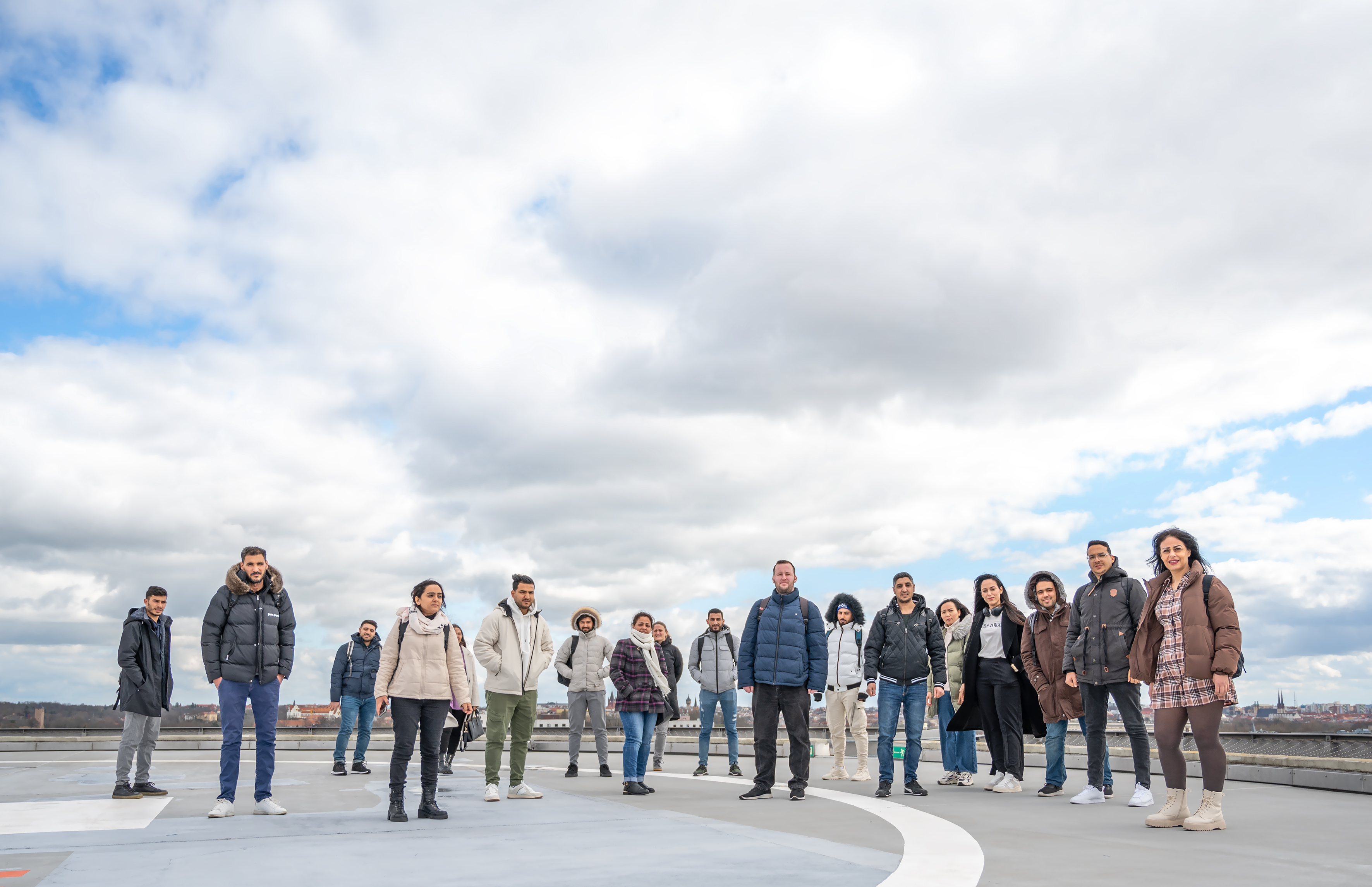 Eine Gruppe junger Menschen steht auf einem Hubschrauberlandeplatz, darüber ist der bewölkte Himmel zu sehen.