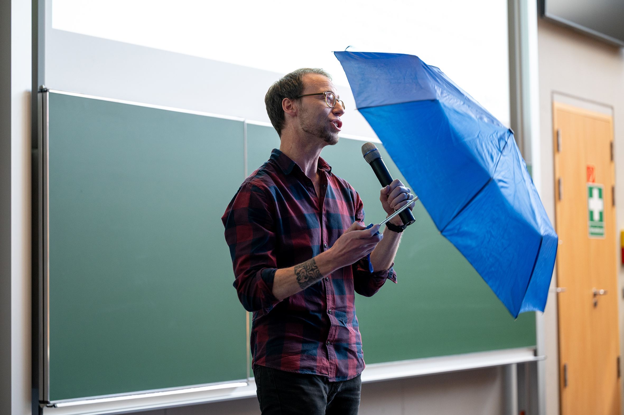Ein Mann hält ein Mikrofon in der linken und einen aufgespannten blauen Regenschirm in der rechten Hand.