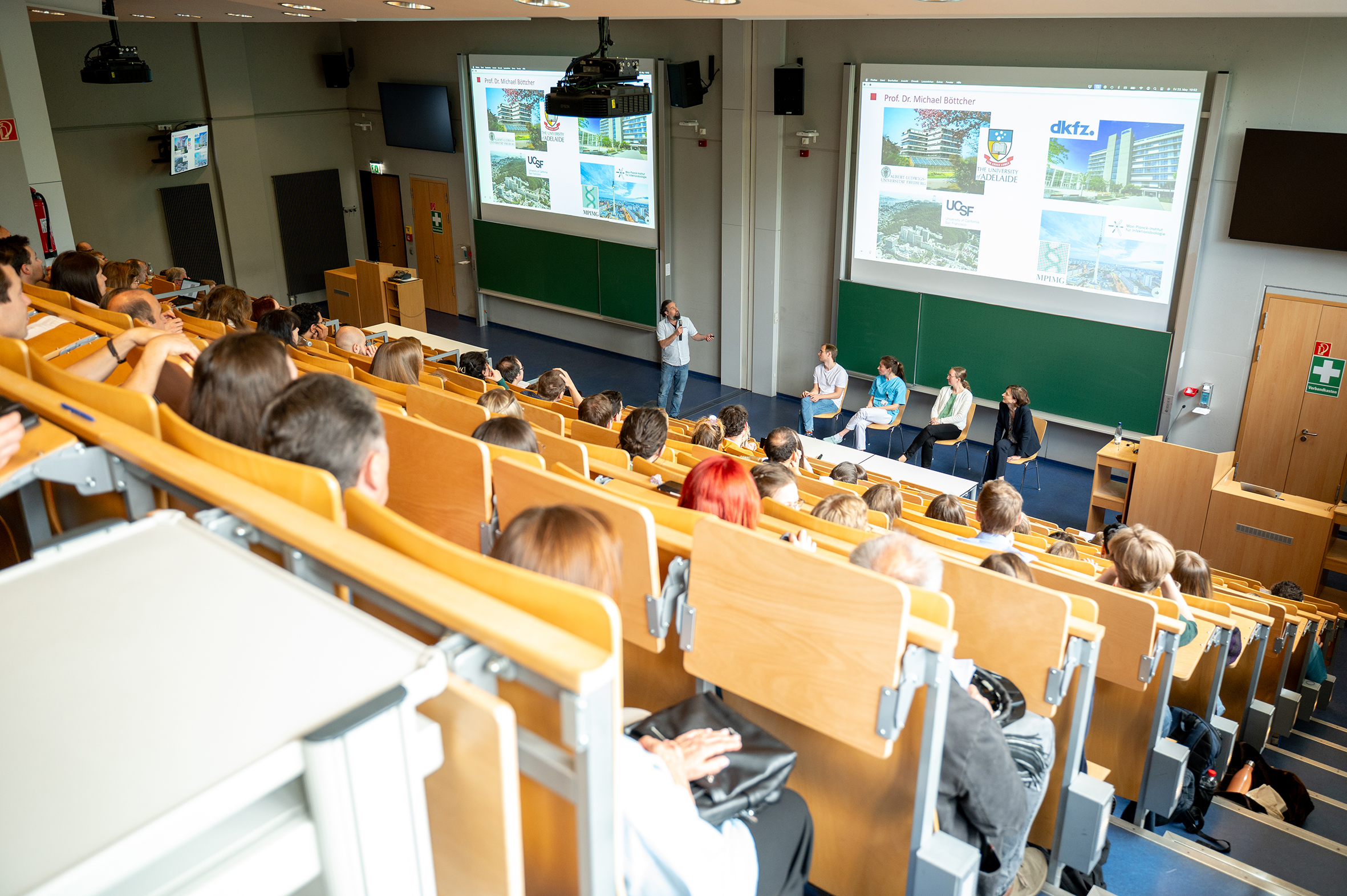 Ein Blick in einen vollen Hörsaal. Vier Personen sitzen vor der Projektionsfläche und hören einer fünften Person zu, die auf die gezeigten Bilder deutet.