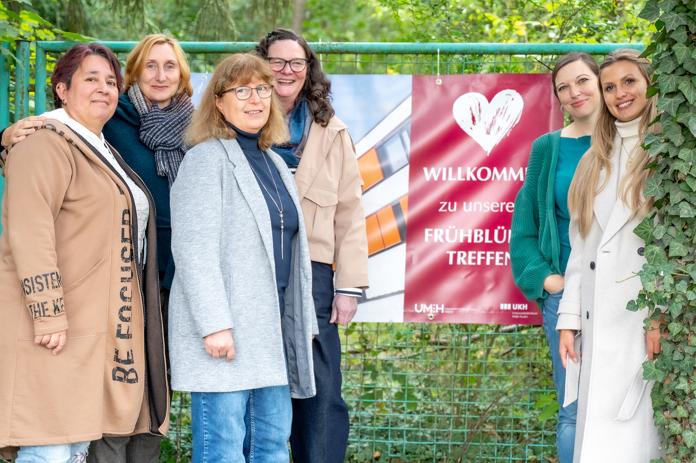 Sechs Erwachsene stehen nebeneinander vor einem grünen Zaun. Sie lächeln freundlich in die Kamera. Neben ihnen hängt ein rotes Banner mit einem weißen Herz und dem Text „Willkommen zu unserem Frühblüher-Treffen“. Im Hintergrund sind Bäume und grüne Pflanzen zu sehen, was auf eine Veranstaltung im Freien hinweist.