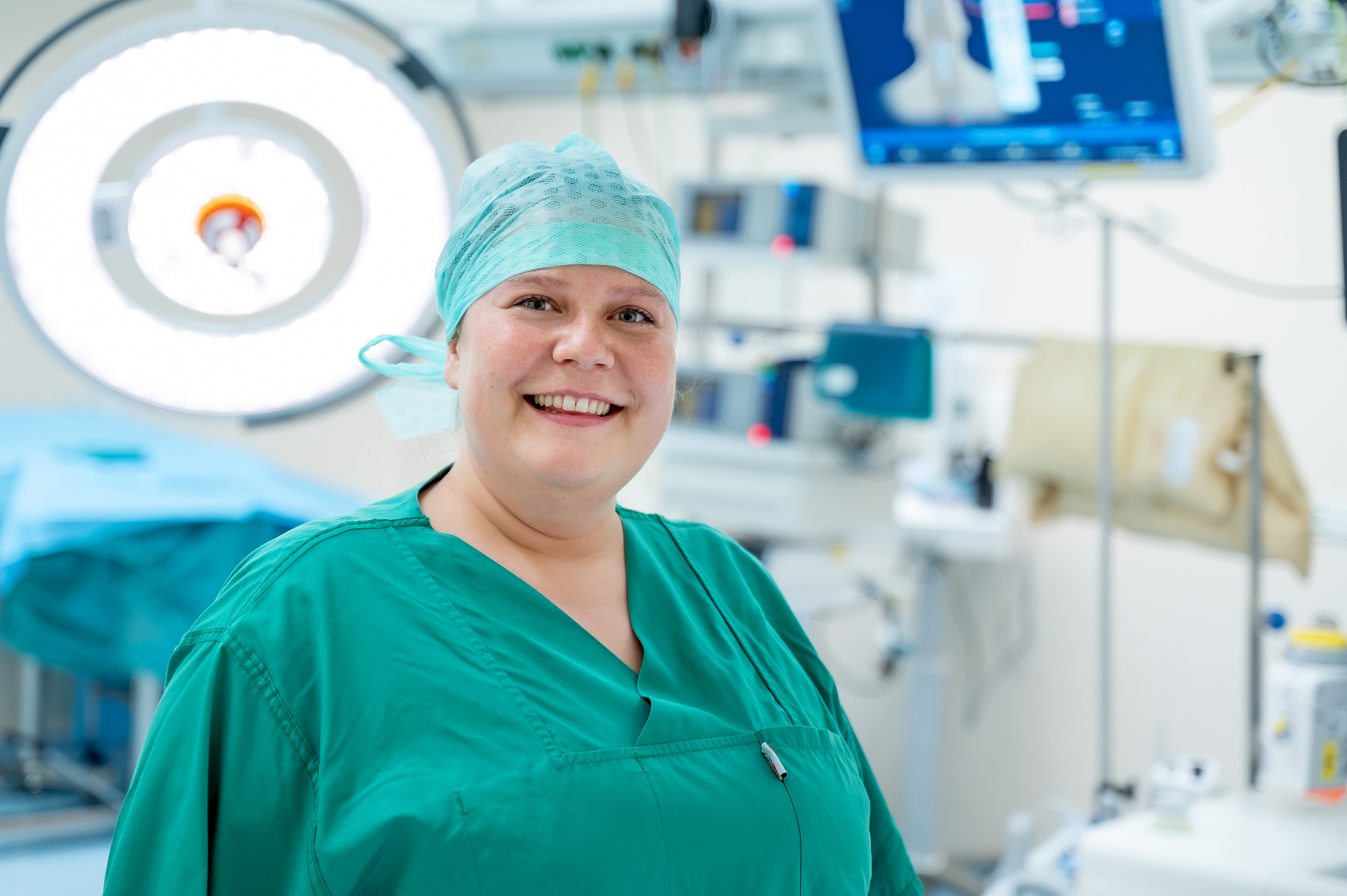 Porträtfoto einer Frau in grüner medizinischer Arbeitskleidung. Im Hintergrund ist ein OP-Saal zu sehen.