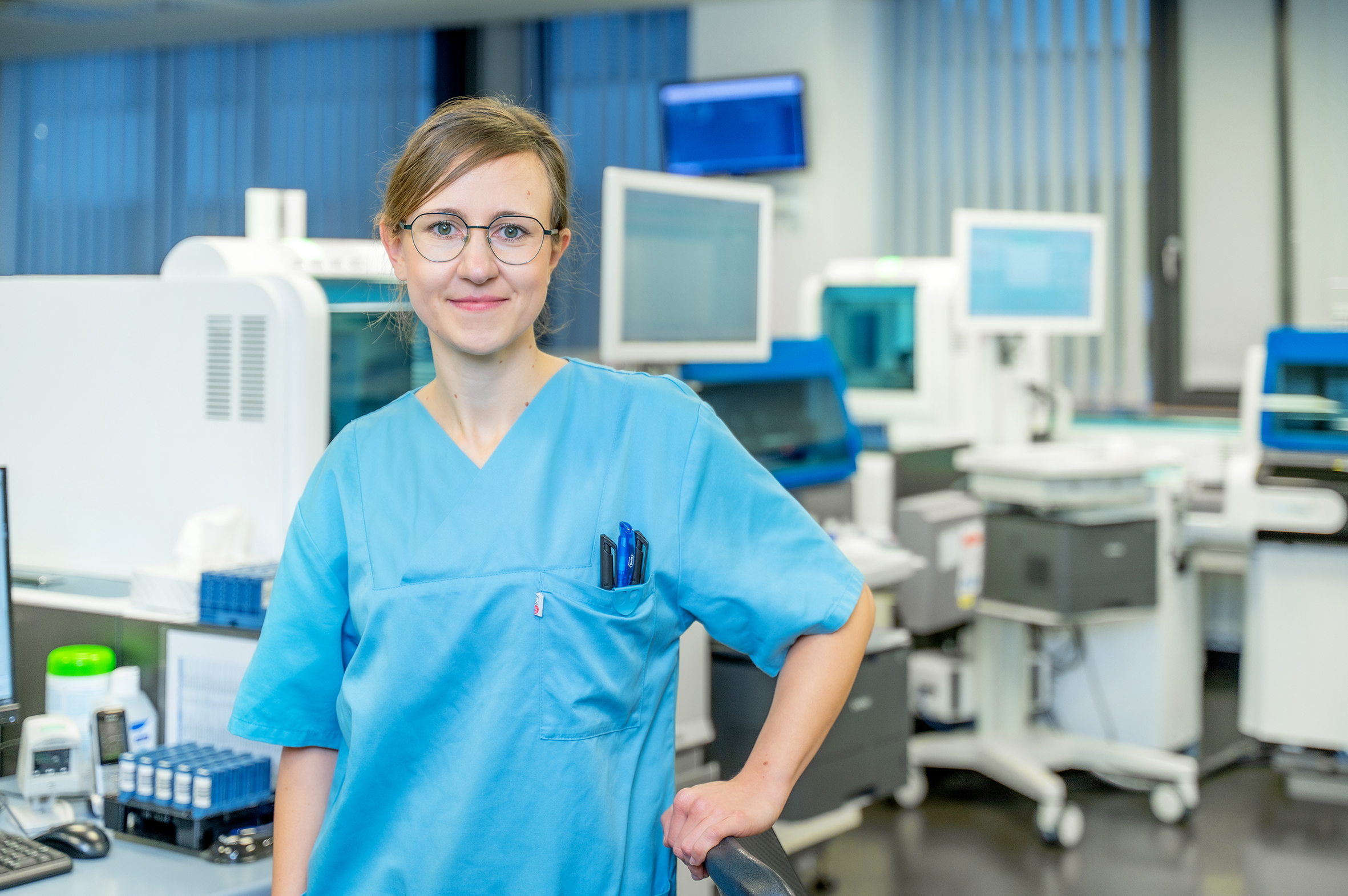 Porträtfoto einer Frau in blauer medizinischer Arbeitskleidung. Im Hintergrund ist ein Großlabor zu sehen.