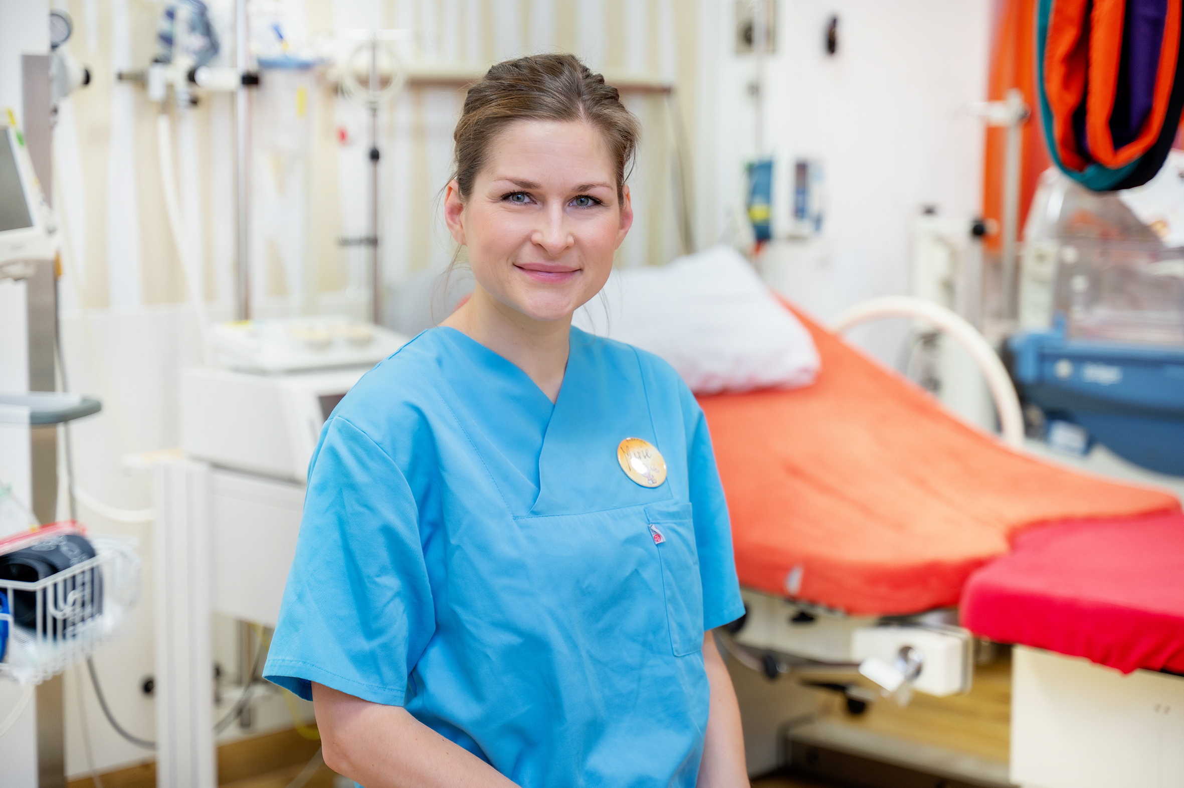 : Porträtfoto einer Frau in blauer medizinischer Arbeitskleidung. Im Hintergrund ist ein Kreißsaal zu sehen.