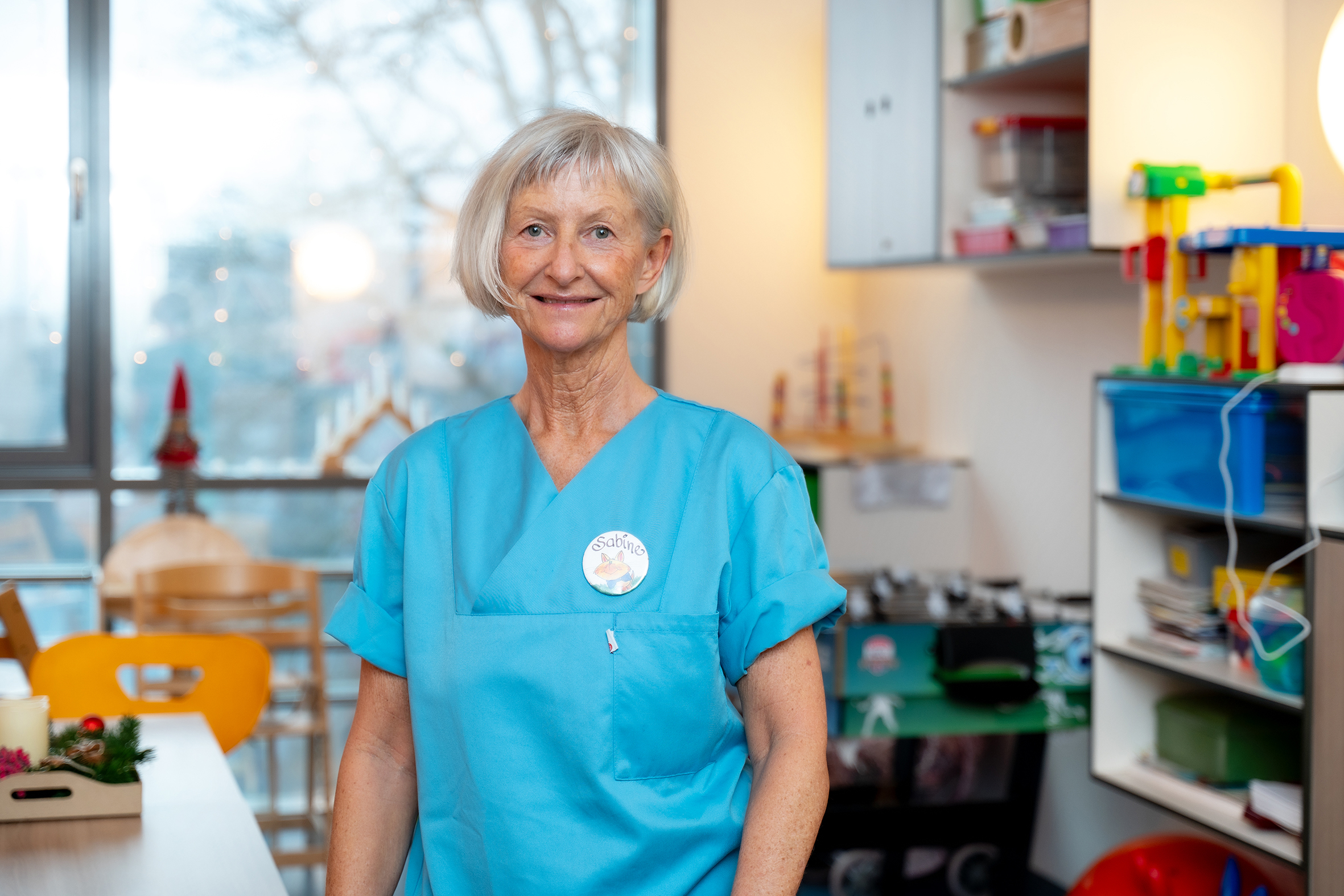 Porträtfoto einer Frau in blauer medizinischer Arbeitskleidung. Im Hintergrund ist ein Spielzimmer einer Krankenhausstation zu sehen.