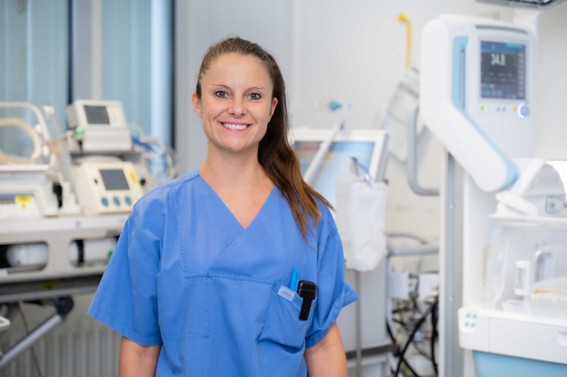 Porträtfoto einer Frau in blauer medizinischer Arbeitskleidung. Im Hintergrund ist ein Intensivkrankenzimmer zu sehen.
