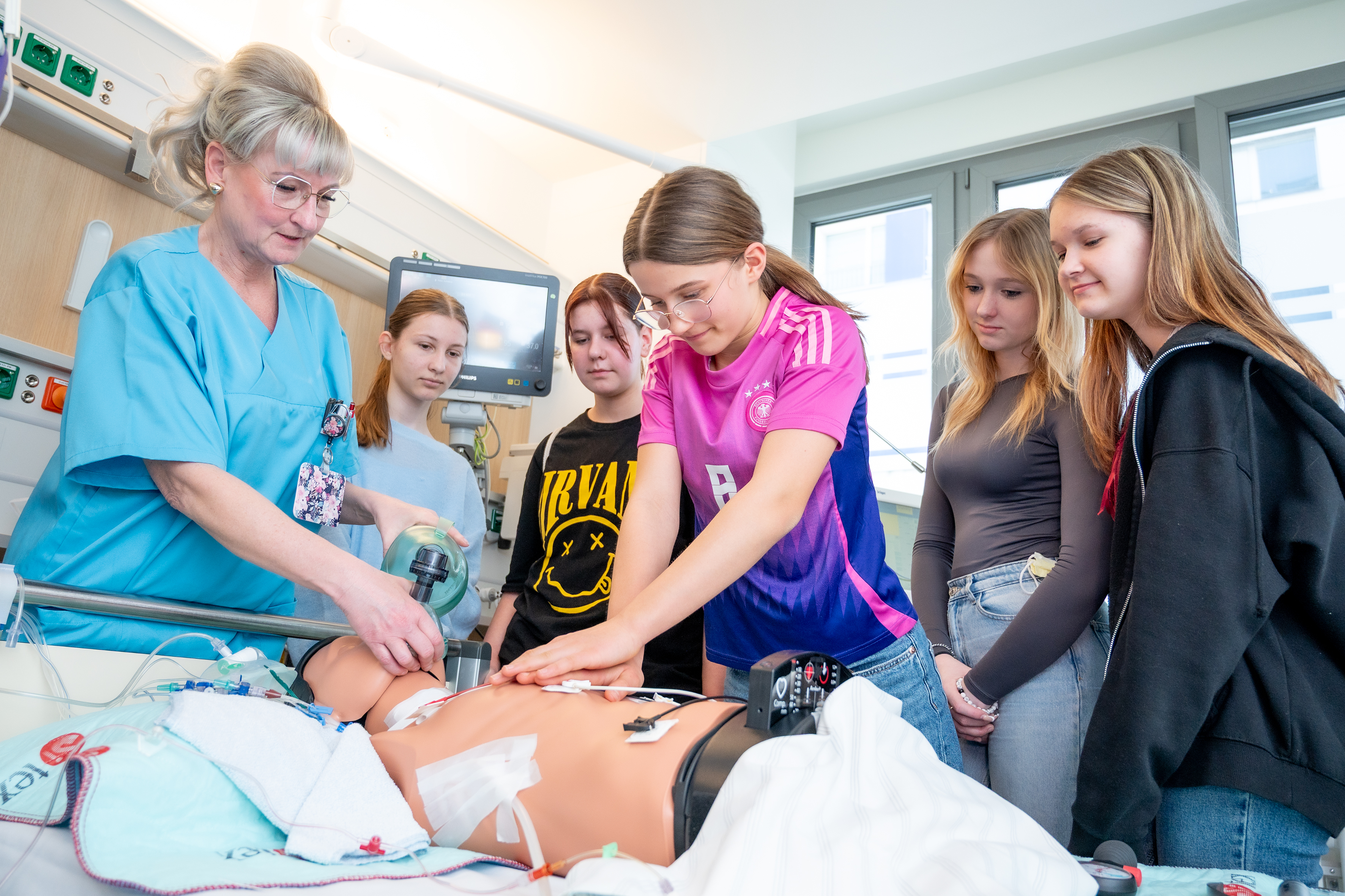 Eine Frau (links) in medizinischer Arbeitskleidung (türkis) leitet eine Gruppe von 4 Mädchen in der Wiederbelebung an einer Übungspuppe an.
