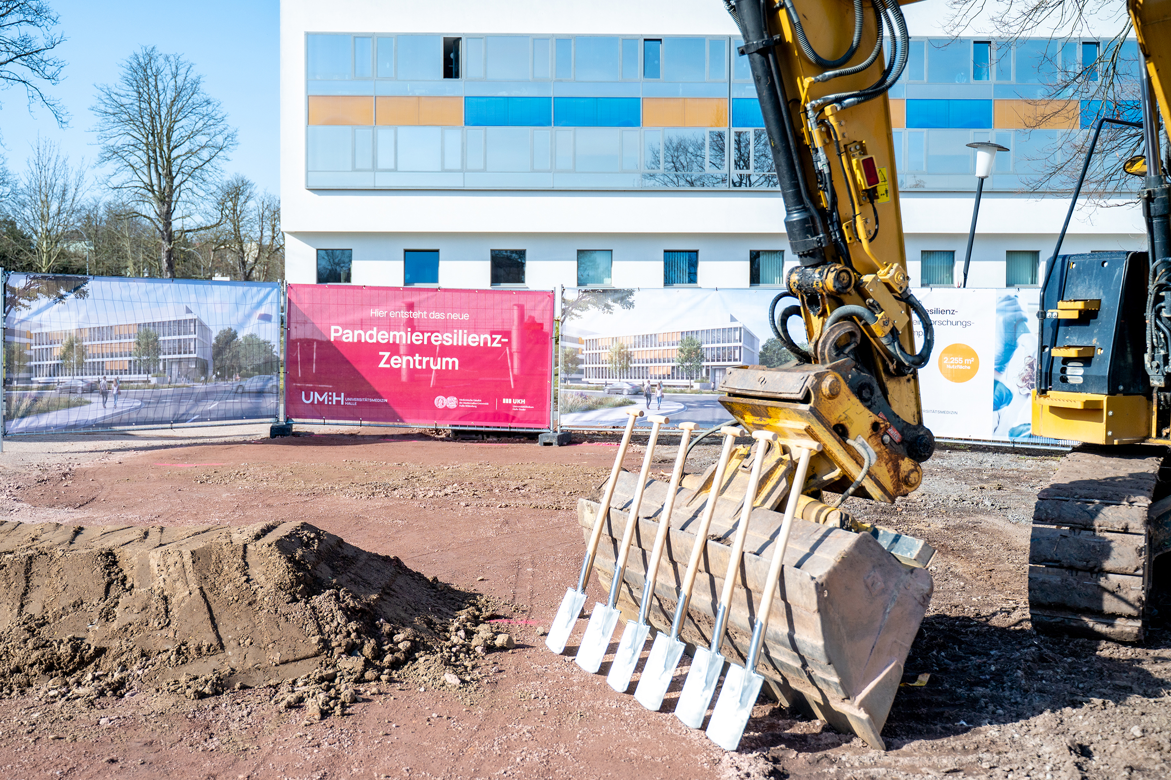 6 Spaten an einen Bagger gelehnt. Im Hintergrund auf einer Bauzaunplane steht Pandemieresilienzzentrum