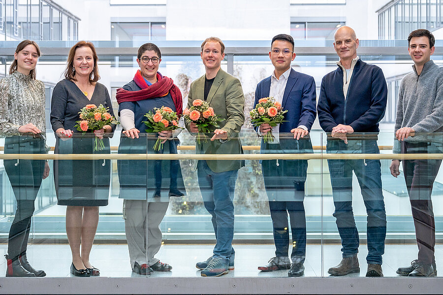Sieben Personen stehen vor einem gläsernen Geländer und lächeln in die Kamera. Vier von ihnen halten einen Blumenstrauß.  Sieben Personen stehen vor einem gläsernen Geländer und lächeln in die Kamera. Vier von ihnen halten einen Blumenstrauß.