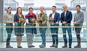 Sieben Personen stehen vor einem gläsernen Geländer und lächeln in die Kamera. Vier von ihnen halten einen Blumenstrauß.