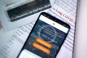 Ein Handy mit der Webseite von DigiHero auf dem Display liegt auf einem Papierhaufen. Auf dem Papiertext ist "Long COVID" rot hervorgehoben. Im oberen linken Bildrand ist ein Briefumschlag mit einer Einladung zur Studie erkennbar.