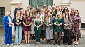 Auf dem Gruppenfoto sind 21 Personen zu sehen. Die 16 anwesenden Absolventinnen des Studiengangs Hebammenwissenschaft halten jeweils einen kleinen Blumenstrauß in den Händen. Auf dem Bild sind außerdem Lehrende und die Studiengangsleitung zu sehen.
