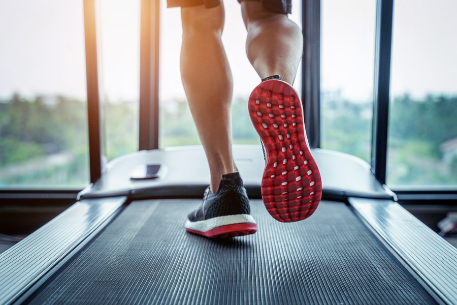 zwei Füße in Turnschuhen mit roter Sohle auf einem Laufband in Bewegung. Das Laufband steht vor einer Fensterfront mit Blick in die Natur.  zwei Füße in Turnschuhen mit roter Sohle auf einem Laufband in Bewegung. Das Laufband steht vor einer Fensterfront mit Blick in die Natur.