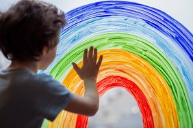 Ein Kleinkind ist von hinten zu sehen. Es legt seine Hand an eine Scheibe auf die ein Regenbogen gemalt ist