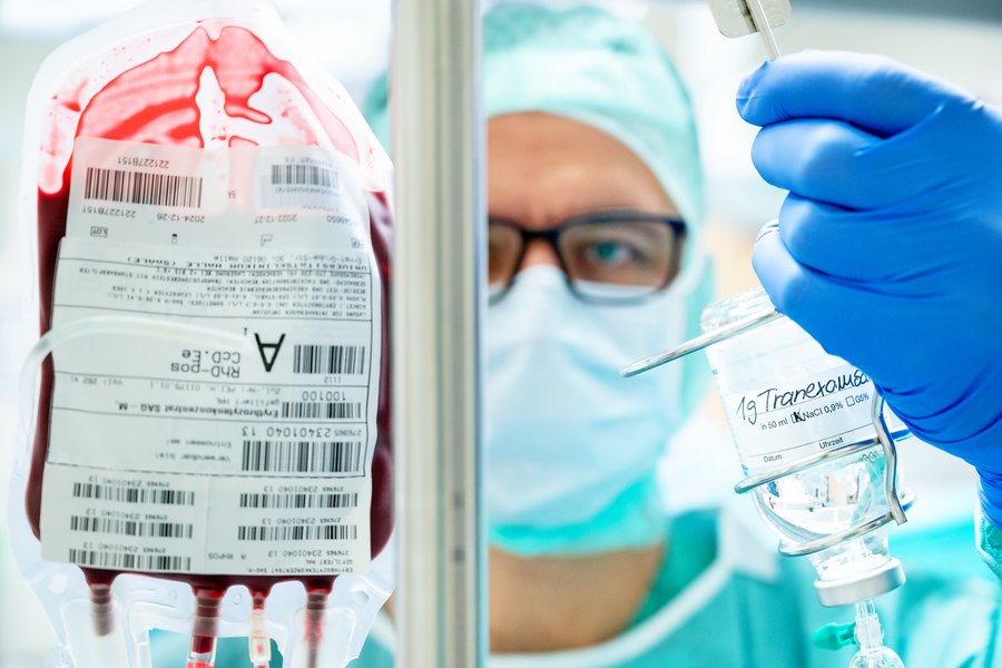 Prof. Ulrich Ronellenfitsch steht vor einem Ständer mit einer Bluttransfusion und blickt in die Kamera. In seiner linken Hand hält er ein Glasfläschchen mit einer klaren Flüssigkeit und der Beschriftung "1g Tranexamsäure".  Prof. Ulrich Ronellenfitsch steht vor einem Ständer mit einer Bluttransfusion und blickt in die Kamera. In seiner linken Hand hält er ein Glasfläschchen mit einer klaren Flüssigkeit und der Beschriftung "1g Tranexamsäure".