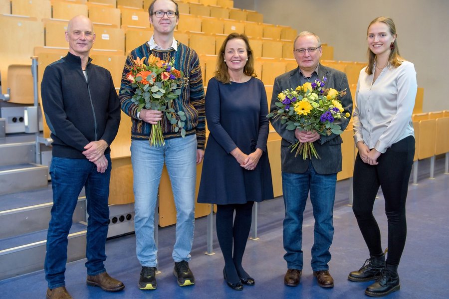 Fünf Personen stehen nebeneinander in einem großen Hörsaal. Zwei Männer halten Blumensträuße.  Fünf Personen stehen nebeneinander in einem großen Hörsaal. Zwei Männer halten Blumensträuße.