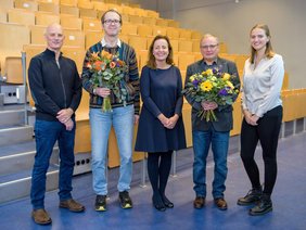 Fünf Personen stehen nebeneinander in einem großen Hörsaal. Zwei Männer halten Blumensträuße.