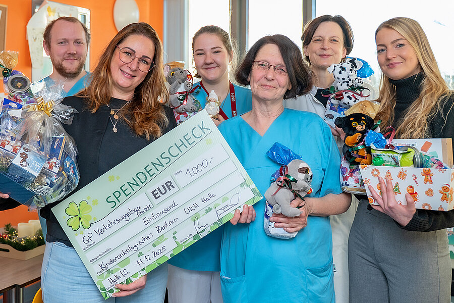Sechs Personen stehen im Aufenthaltsraum einer Kinderstation im UKH. Einige Personen halten einen Spendenscheck über 1000 Euro und Geschenke in den Händen.  Sechs Personen stehen im Aufenthaltsraum einer Kinderstation im UKH. Einige Personen halten einen Spendenscheck über 1000 Euro und Geschenke in den Händen.