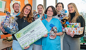 Sechs Personen stehen im Aufenthaltsraum einer Kinderstation im UKH. Einige Personen halten einen Spendenscheck über 1000 Euro und Geschenke in den Händen.