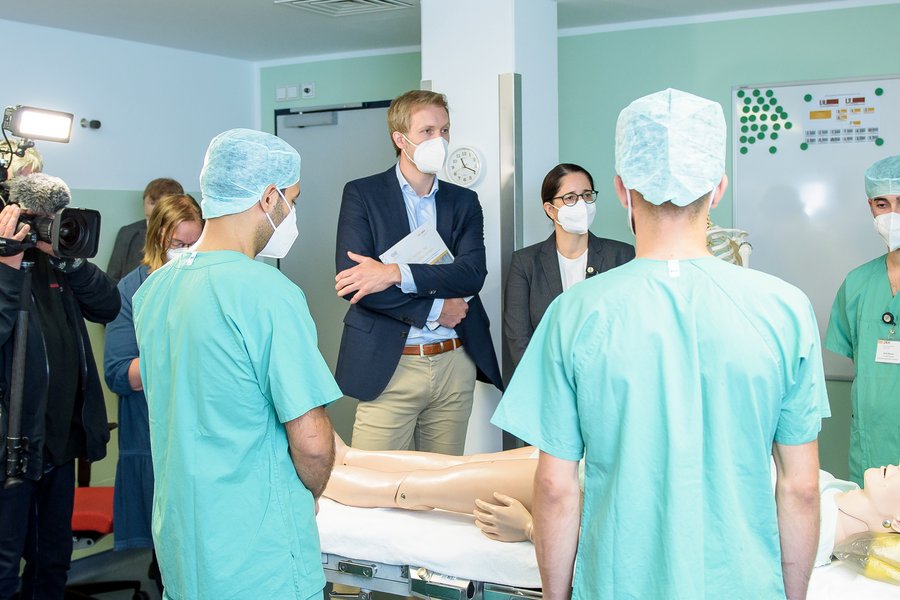 Menschen in ziviler und medizinischer Arbeitsschutzkleidung stehen um ein Krankenbett, in dem eine Patientenpuppe liegt.  Menschen in ziviler und medizinischer Arbeitsschutzkleidung stehen um ein Krankenbett, in dem eine Patientenpuppe liegt.