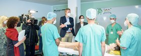 Menschen in ziviler und medizinischer Arbeitsschutzkleidung stehen um ein Krankenbett, in dem eine Patientenpuppe liegt.