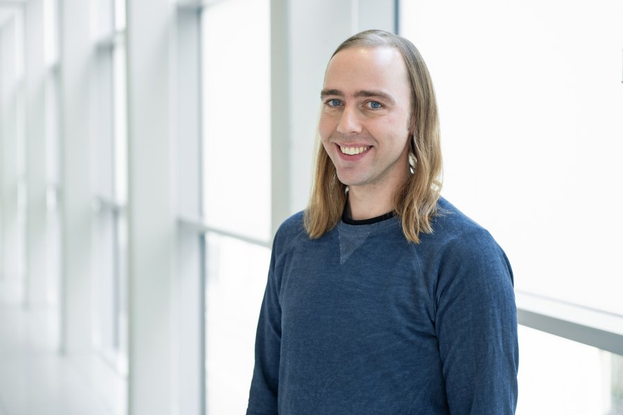 Mann mit langen Haaren steht in einem Glasgang und blickt in die Kamera. Er trägt ein dunkles Shirt.   Mann mit langen Haaren steht in einem Glasgang und blickt in die Kamera. Er trägt ein dunkles Shirt. 