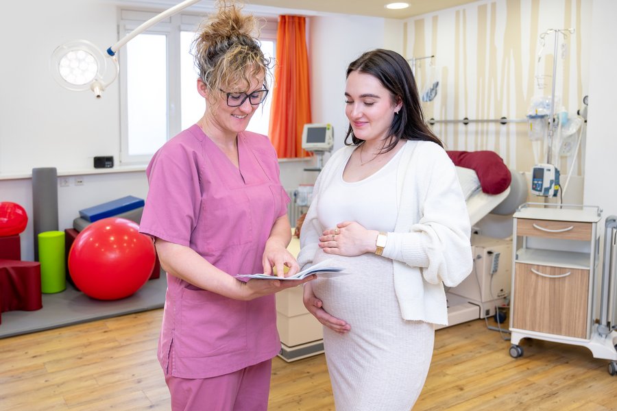 Eine Hebamme in rosa Arbeitskleidung und eine schwangere Frau schauen sich gemeinsam einen Mutterpass an. Sie stehen in einem hellen, modern eingerichteten Kreißsaal. Im Hintergrund sind viele Utensilien sowie ein Kreißbett und technische Geräte zu sehen.  Eine Hebamme in rosa Arbeitskleidung und eine schwangere Frau schauen sich gemeinsam einen Mutterpass an. Sie stehen in einem hellen, modern eingerichteten Kreißsaal. Im Hintergrund sind viele Utensilien sowie ein Kreißbett und technische Geräte zu sehen.