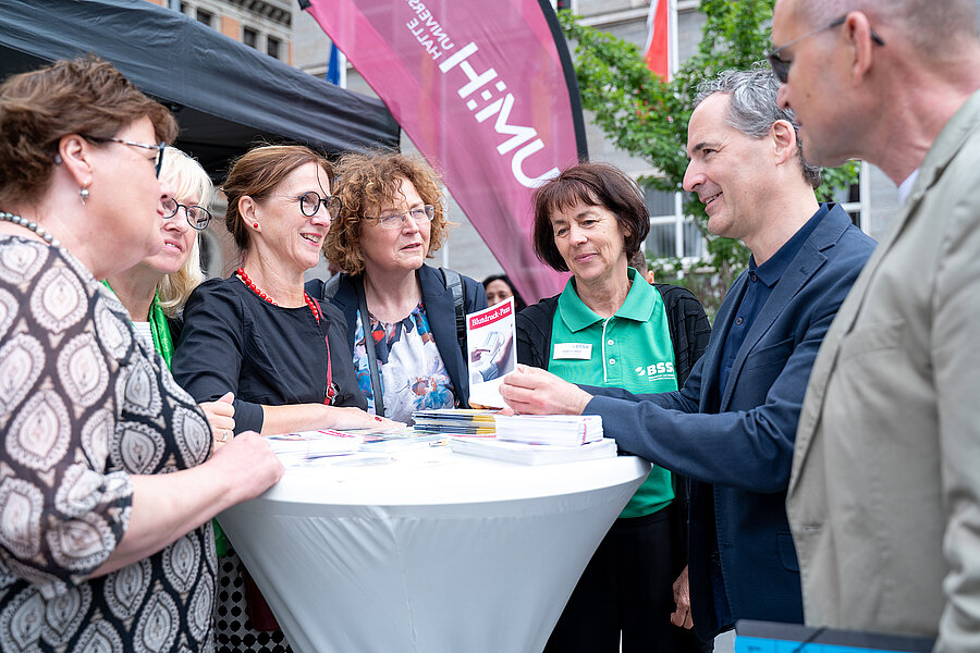 Sieben Personen stehen auf dem Marktplatz in Halle (Saale) um einen Stehtisch herum und unterhalten sich. Im Hintergrund befindet sich eine Beachflag der Universitätsmedizin Halle.  Sieben Personen stehen auf dem Marktplatz in Halle (Saale) um einen Stehtisch herum und unterhalten sich. Im Hintergrund befindet sich eine Beachflag der Universitätsmedizin Halle.