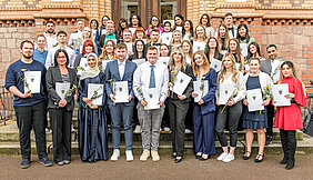 Eine große Gruppe von Personen posiert für ein Gruppenfoto. Die meisten Personen sind festlich gekleidet. Sie halten ihre Zeugnisse und Blumen in den Händen. 