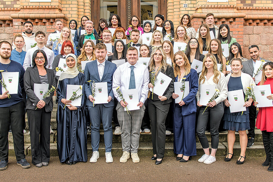 Eine große Gruppe von Personen posiert für ein Gruppenfoto. Die meisten Personen sind festlich gekleidet. Sie halten ihre Zeugnisse und Blumen in den Händen.   Eine große Gruppe von Personen posiert für ein Gruppenfoto. Die meisten Personen sind festlich gekleidet. Sie halten ihre Zeugnisse und Blumen in den Händen. 