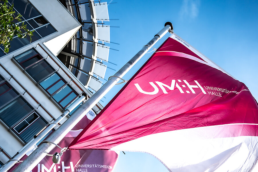 magentafarbene Fahne mit Aufdruck UMH flattert vor blauem Himmel und einer angeschnittenen Fassade im Wind.  magentafarbene Fahne mit Aufdruck UMH flattert vor blauem Himmel und einer angeschnittenen Fassade im Wind.