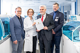 drei Männer und eine Frau stehen in einem Labor mit Laborgeräten im Hintergrund. Es wird ein Fördermittelbescheid übergeben.