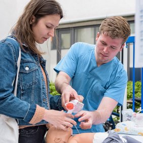 Eine Person in Klinikkleidung beatmet eine Übungspuppe eines Säuglings. Eine weitere Person mit Jeansjacke und Beutel assistiert dabei.