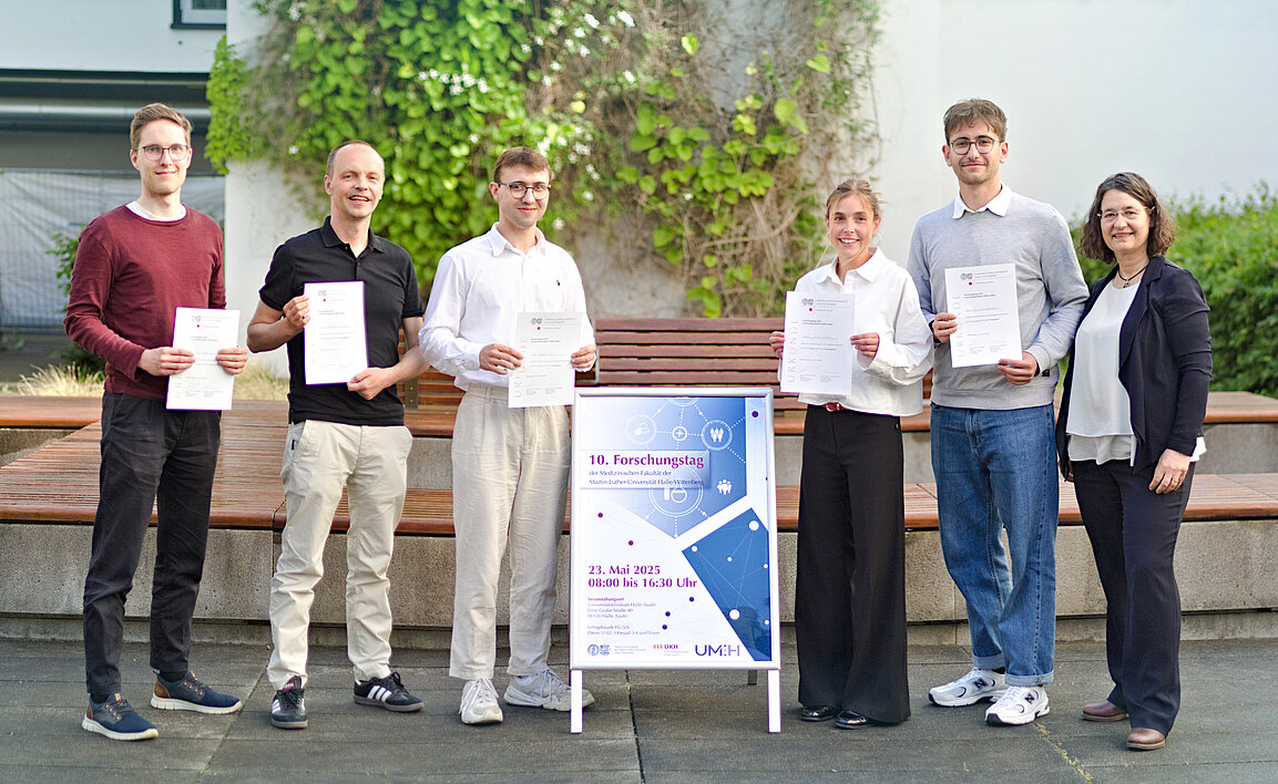 Sechs Personen stehen in einem Hof und lächeln in die Kamera. In deren Mitte befindet sich ein Plakataufsteller mit der Aufschrift „10. Forschungstag“. Fünf der Personen halten eine Urkunde in den Händen. Im Hintergrund sind eine Sitzgruppe und Pflanzen an einer Hauswand unscharf zu erkennen.