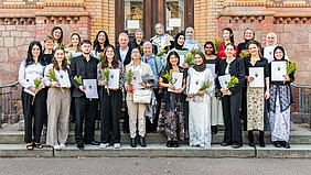 Die Absolvent:innen stehen vor einem Gebäude und schauen in die Kamera. Sie halten ihre Zeugnisse und Blumen in den Händen.