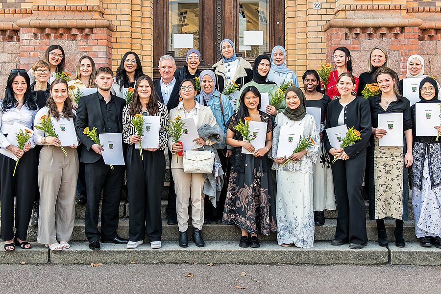 Die Absolvent:innen stehen vor einem Gebäude und schauen in die Kamera. Sie halten ihre Zeugnisse und Blumen in den Händen.  Die Absolvent:innen stehen vor einem Gebäude und schauen in die Kamera. Sie halten ihre Zeugnisse und Blumen in den Händen.