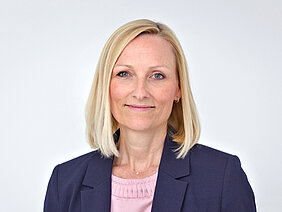 Zu sehen ist eine Frau mit mittellangen blonden Haaren, einer Goldkette und einem Blazer, die direkt in die Kamera schaut. Der Hintergrund ist neutral gehalten.