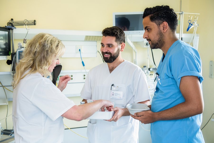 Zwei Männer und eine Frau in weißen und blauen Kassaks stehen zusammen in einem Patient:innenzimmer. Die Frau erklärt dem Mann in der Mitte etwas zu medizinischem Material auf einem Tablett. Der andere Mann schaut beiden zu.  Zwei Männer und eine Frau in weißen und blauen Kassaks stehen zusammen in einem Patient:innenzimmer. Die Frau erklärt dem Mann in der Mitte etwas zu medizinischem Material auf einem Tablett. Der andere Mann schaut beiden zu.