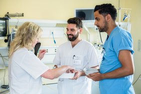 Zwei Männer und eine Frau in weißen und blauen Kassaks stehen zusammen in einem Patient:innenzimmer. Die Frau erklärt dem Mann in der Mitte etwas zu medizinischem Material auf einem Tablett. Der andere Mann schaut beiden zu.