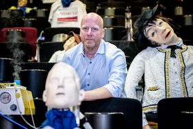 Zu sehen ist ein Zuschauerraum mit Stuhlreihen in einem Theater. auf den Stühlen sitzen Puppen, sowohl vom Theater als auch spezielle Messpuppen. Im Zentrum des Bildes sitzt Dr. Stefan Moritz. Am linken Bildrand ist ein Apparat zu sehen, der nach oben Dampf ausstößt.