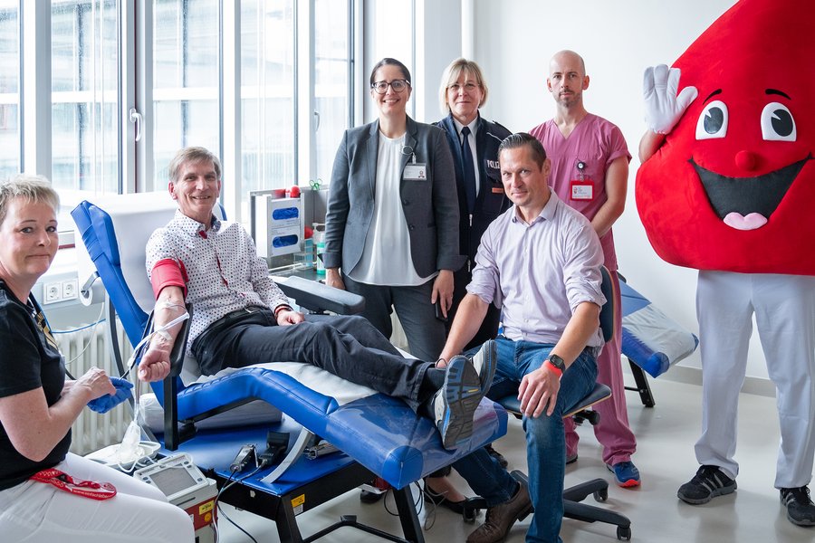 Gruppenbild beim Blutspendedienst: Ein Mann liegt auf einer blauen Liege, er hält den rechten Arm nach unten ausgestreckt, daran hängt eine Blutspendenadel. Neben ihm stehen und sitzen drei Frauen und zwei Männer sowie ein Mensch im Maskottchenkostüm eines freundlich schauen-den Blutstropfens. Eine Frau trägt eine Polizeiuniform, ein Mann beerenfarbene medizinische Arbeitskleidung. Die anderen sind in zivil gekleidet.  Gruppenbild beim Blutspendedienst: Ein Mann liegt auf einer blauen Liege, er hält den rechten Arm nach unten ausgestreckt, daran hängt eine Blutspendenadel. Neben ihm stehen und sitzen drei Frauen und zwei Männer sowie ein Mensch im Maskottchenkostüm eines freundlich schauen-den Blutstropfens. Eine Frau trägt eine Polizeiuniform, ein Mann beerenfarbene medizinische Arbeitskleidung. Die anderen sind in zivil gekleidet.