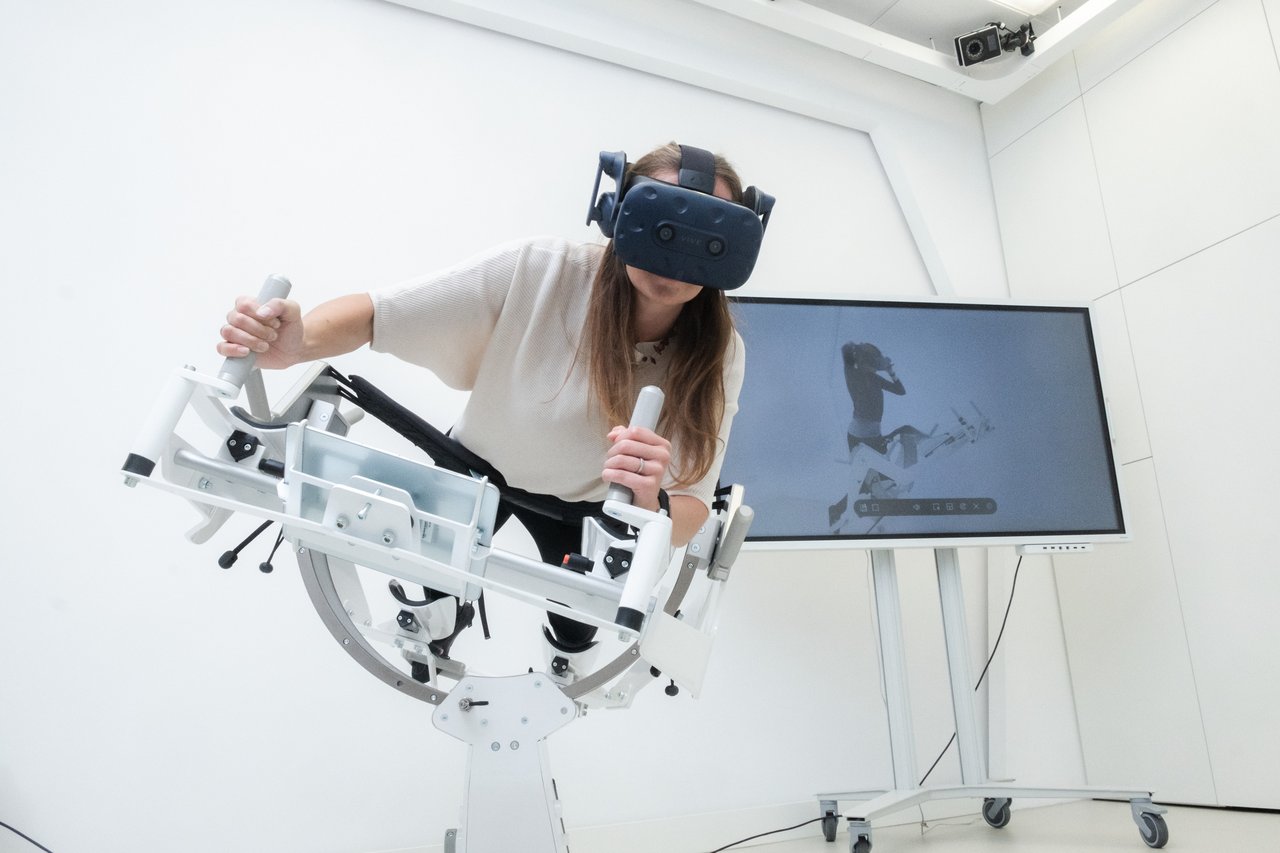 VR-Fitnessgerät „Icaros Health“: Futuristisch aussehendes Gerät, auf dem eine Frau mit VR-Brille liegt und das sie mit ihren Händen steuert, im Hintergrund ist ein Monitor zu sehen.
