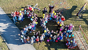 Aufnahme aus der Vogelperspektive. Eine Gruppe von Kindern und einigen Erwachsenen posiert in Form einer 20 auf einer Grünfläche.