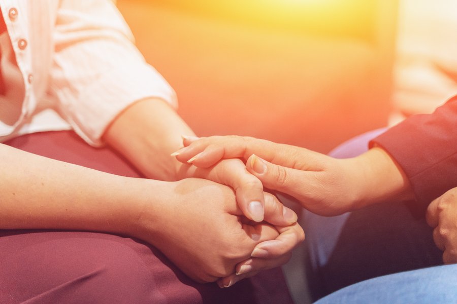 Man sieht die Hände von zwei Menschen. Die eine Person hält beide Hände locker gefaltet im Schoß. Die andere Person hat eine Hand darauf gelegt. Die Geste wirkt vertraut. Im Hintergrund ist gelb-oranges Licht zu sehen.  Man sieht die Hände von zwei Menschen. Die eine Person hält beide Hände locker gefaltet im Schoß. Die andere Person hat eine Hand darauf gelegt. Die Geste wirkt vertraut. Im Hintergrund ist gelb-oranges Licht zu sehen.