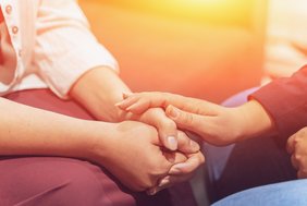 Man sieht die Hände von zwei Menschen. Die eine Person hält beide Hände locker gefaltet im Schoß. Die andere Person hat eine Hand darauf gelegt. Die Geste wirkt vertraut. Im Hintergrund ist gelb-oranges Licht zu sehen.