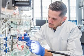 Ein Mann sitzt der Kamera zugewandt an einem Labortisch, im Hintergrund ist ein Fenster zu erkennen. Mit blauen Nitrilhandschuhen ausgestattet arbeitet er an einem Versuchsaufbau, in dem ein kleines Rattenherz zwischen zahlreichen Schläuchen und Becken scheinbar frei schwebt.