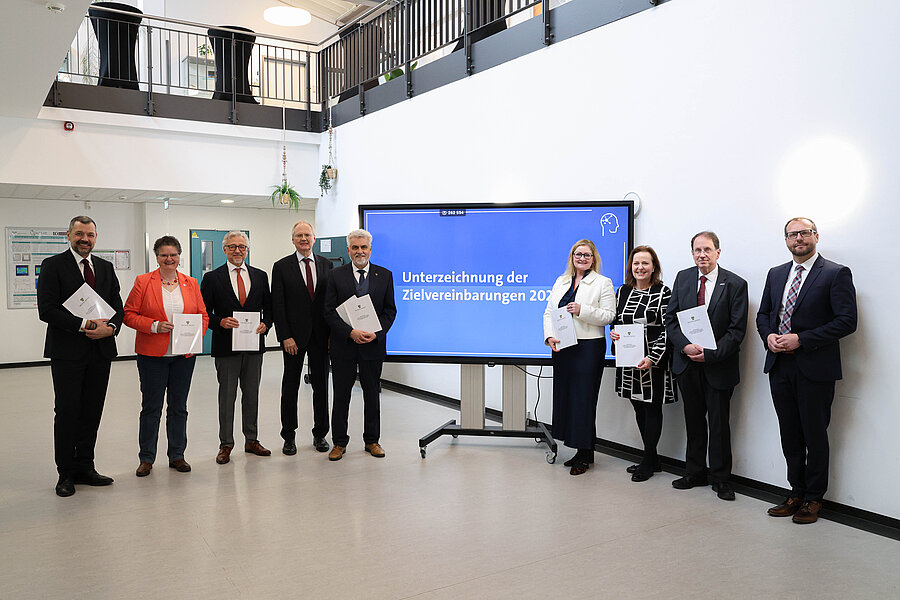 Neun Personen stehen in einer Reihe nebeneinander im Raum des Forschungscampus STIMULATE. In der Mitte der Reihe befindet sich ein großer Bildschirm mit dem Bildtext „Unterzeichnung der Zielvereinbarung“.  Neun Personen stehen in einer Reihe nebeneinander im Raum des Forschungscampus STIMULATE. In der Mitte der Reihe befindet sich ein großer Bildschirm mit dem Bildtext „Unterzeichnung der Zielvereinbarung“.