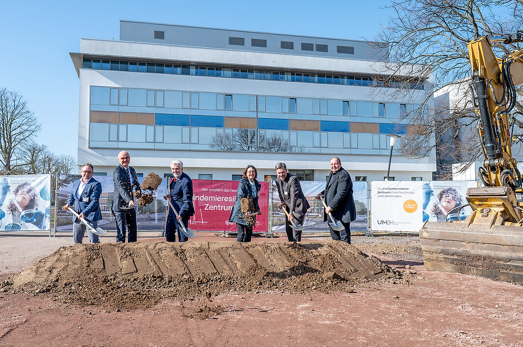 Zu sehen sind 6 Personen. Jede wirft mit einem Spaten Erde aus einem aufgeschütteten Haufen in die Luft. Hinter den Personen ist das LZG des Uni-Klinikums zu sehen, rechts steht ein großer gelber Bagger. An einem Bauzaun zwischen LZG und den Personen sind Banner aufgehängt. Eins ist mit "Pandemieresilienz-Zentrum" in weißer Schrift auf magenta-farbenem Hintergrund bedruckt. Ein weiteres enthält die Visualisierung, wie das Gebäude einmal aussehen soll. Ein drittes erklärt: "Das Pandemieresilienz-Zentrum ist ein Forschungs- und Laborgebäude".  